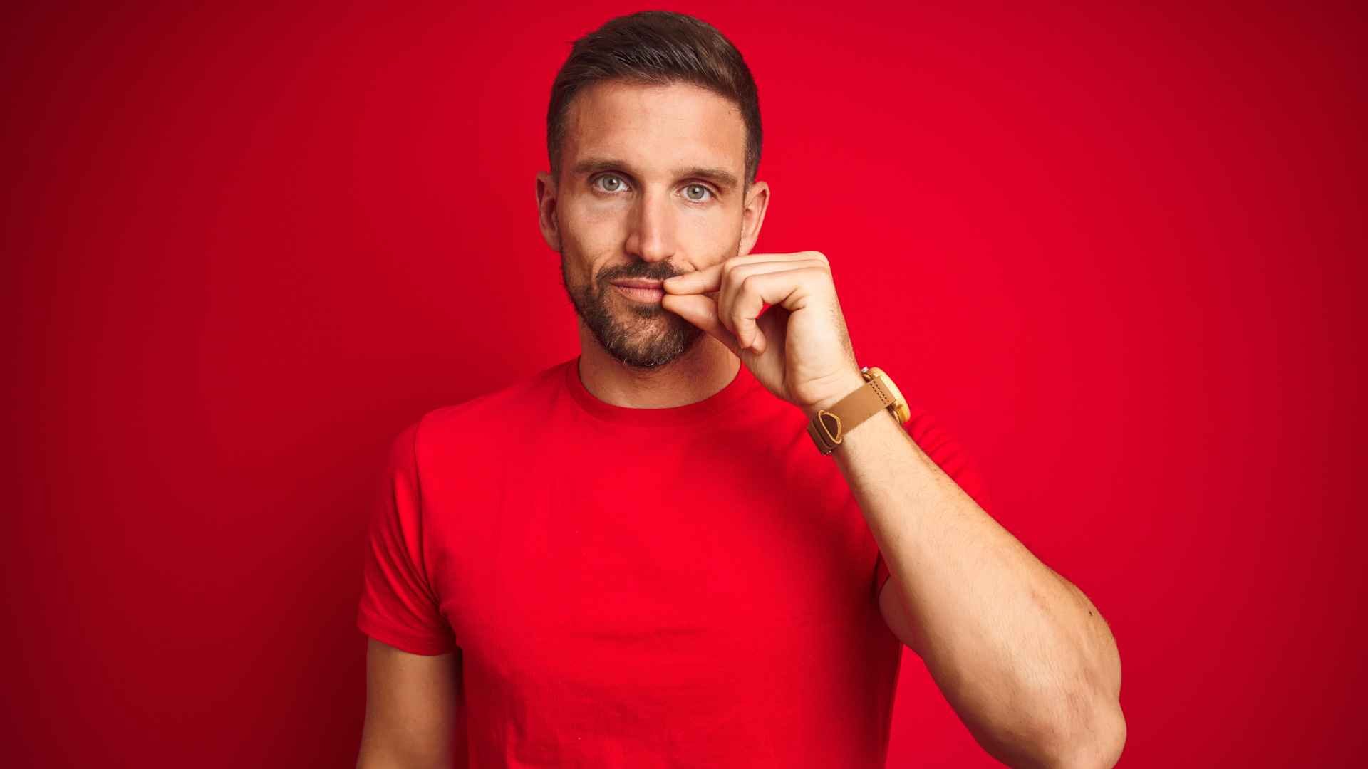Junger Mann in einem roten Shirt vor einem roten Hintergrund, der die erste Regel des Mom Tests beherzigt und seine Lippen wie mit einem Reißverschluss verschließt.