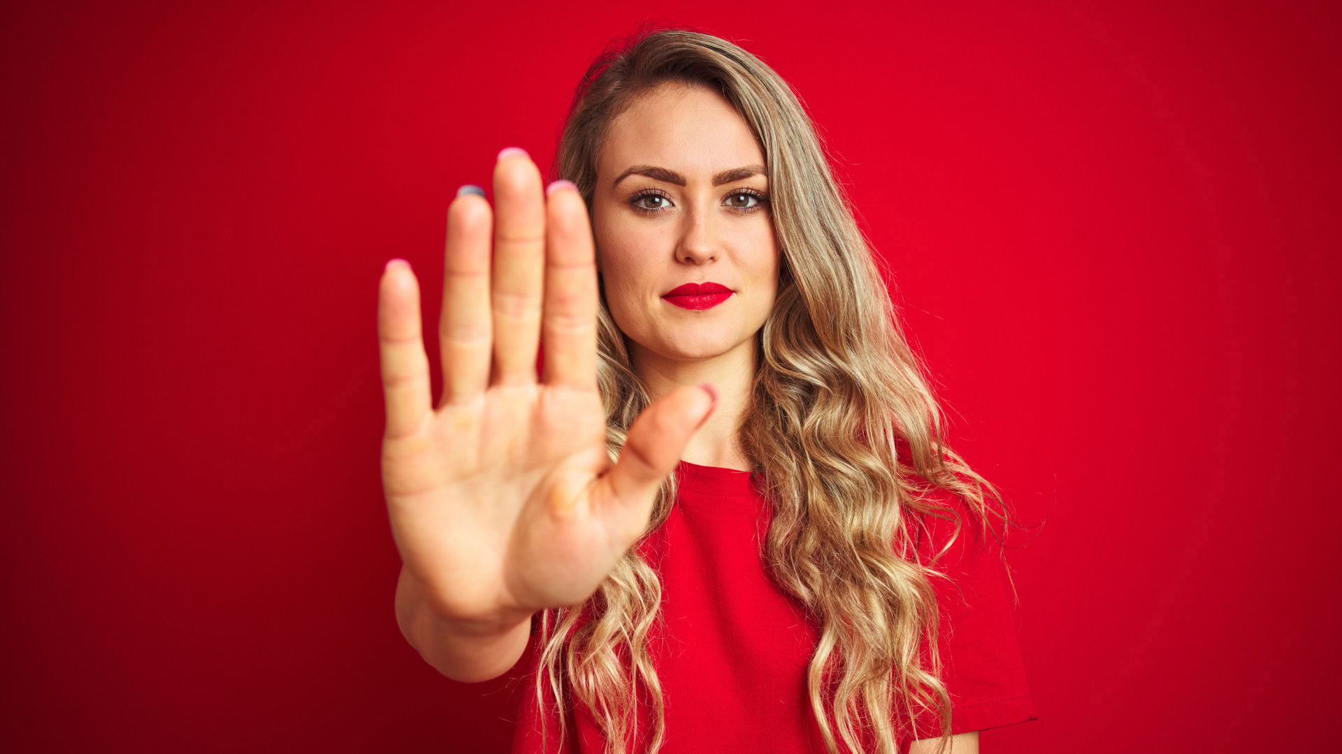 Eine Frau in roter Kleidung steht vor einem roten Hintergrund und streckt die Hand nach vorne, als würde sie eine Grenze setzen. Ihr Gesichtsausdruck ist entschlossen und selbstbewusst. Dieses Bild symbolisiert ablehnende Nichtkunden in der Blue-Ocean-Strategie – Menschen, die sich bereits mit einer Branche auseinandergesetzt haben, sich aber bewusst gegen deren Angebote entschieden haben.
