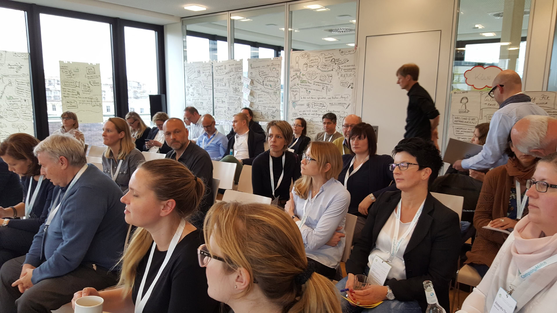 Eine Abschlussrunde bei einem Barcamp mit einer Gruppe von Teilnehmenden, die konzentriert zuhören. Im Hintergrund sind großflächige Sketchnotes mit visuellen Zusammenfassungen der Sessions zu sehen.