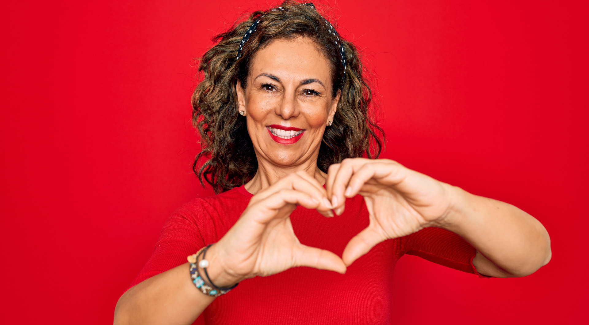 Fröhliche Frau in rotem Shirt vor rotem Hintergrund formt mit den Händen ein Herz und lächelt in die Kamera.