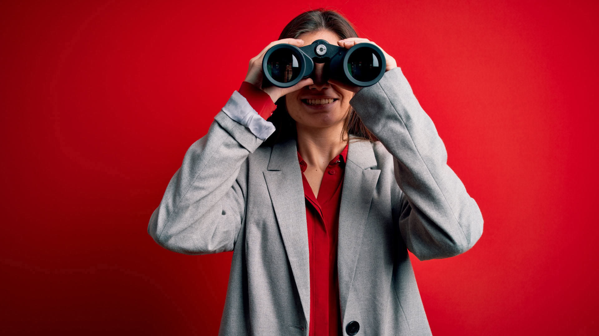 Frau im Business-Outfit blickt durch ein Fernglas vor rotem Hintergrund — symbolisch für die Phase des aktiven Suchens im Switch Interview.