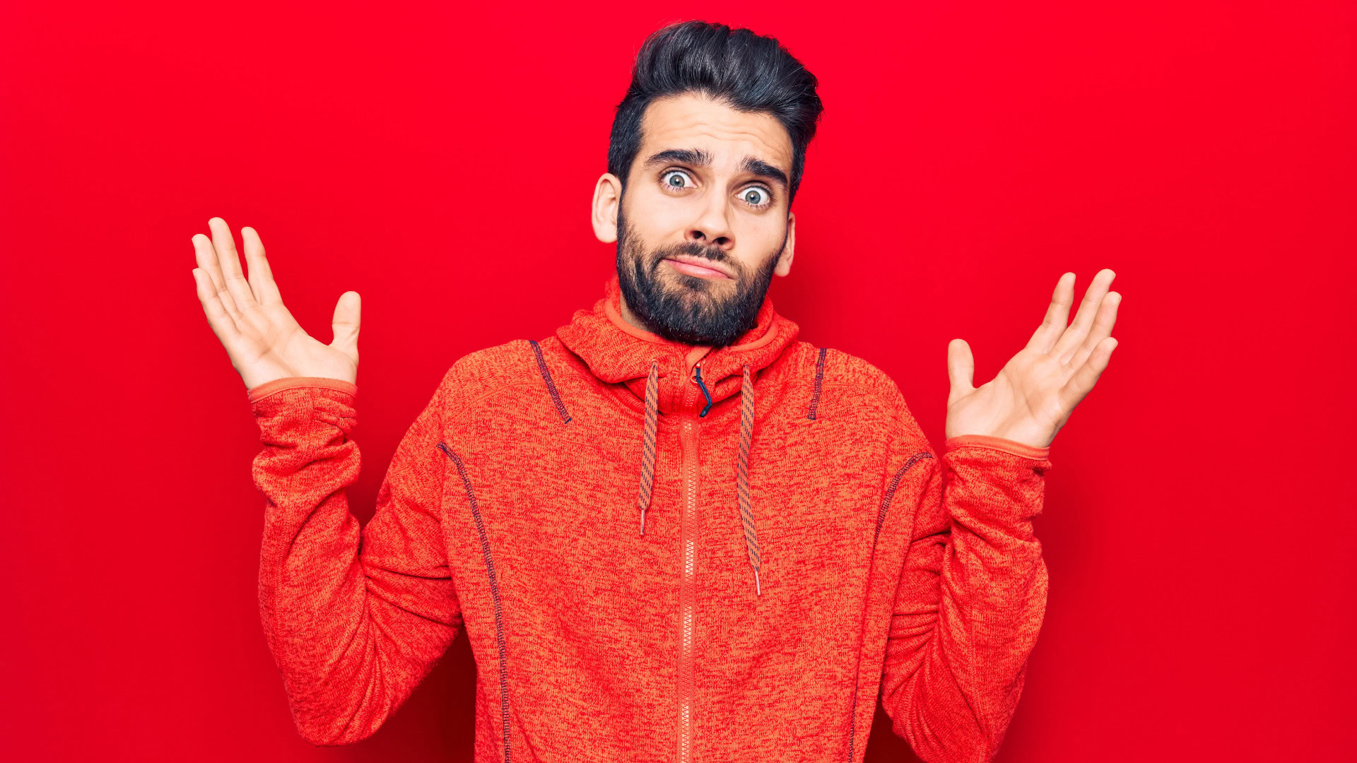 Mann mit verwirrtem Gesichtsausdruck hebt beide Hände vor einem roten Hintergrund, symbolisiert Unsicherheit durch zu viele OKR.