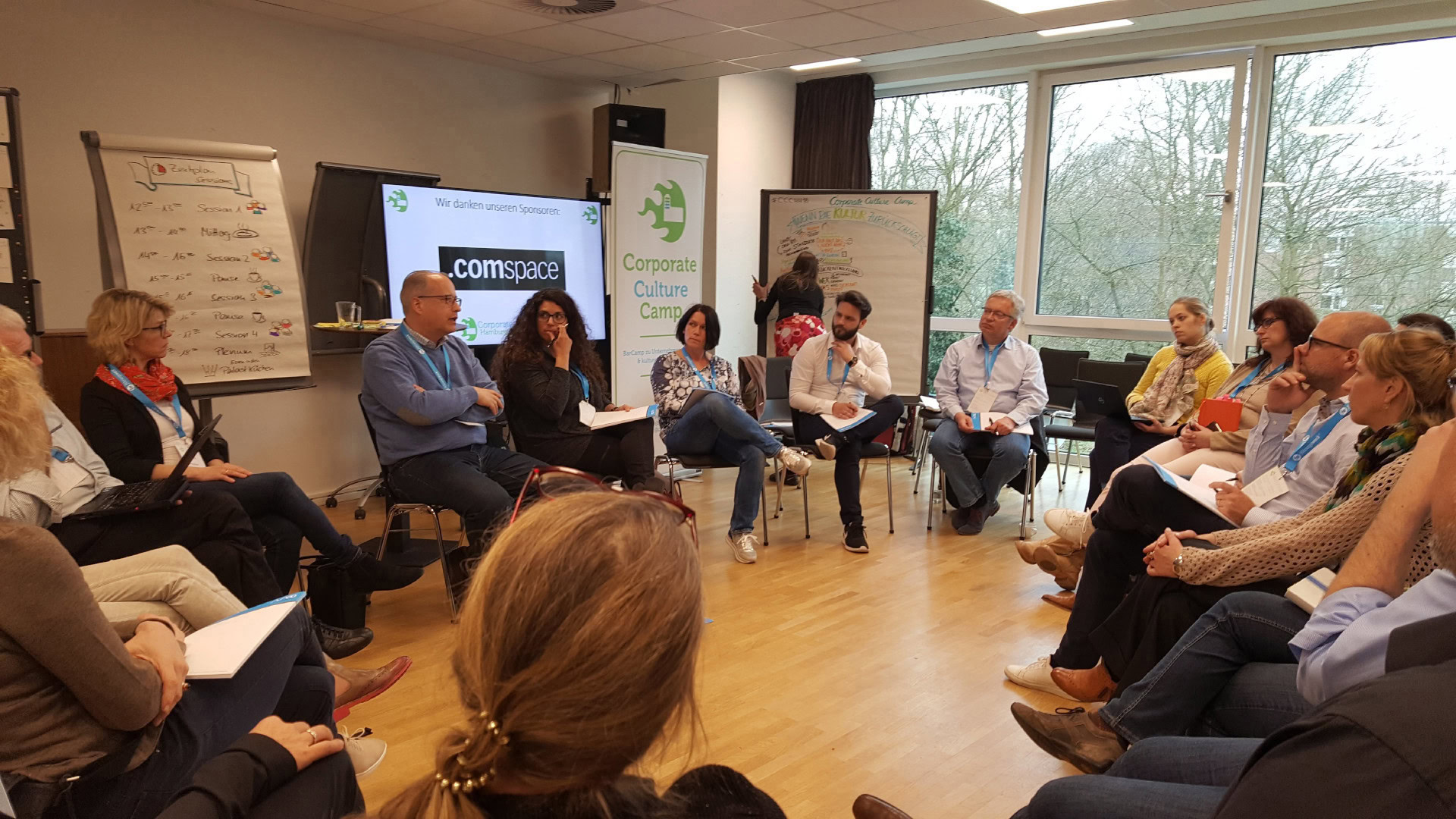 Eine Barcamp-Session in einem lichtdurchfluteten Raum zeigt eine Gruppe von Teilnehmenden, die im Stuhlkreis sitzen und aktiv an der Diskussion teilnehmen. Im Hintergrund ist ein Whiteboard mit einer visuellen Dokumentation zu sehen.