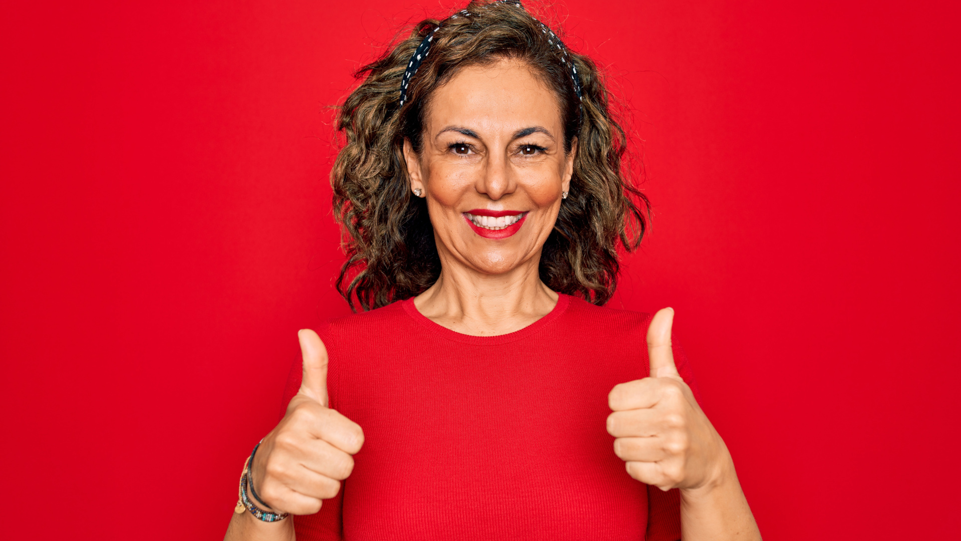 Ältere Frau in rotem Shirt vor rotem Hintergrund. Streckt beide Daumen nach oben, um den bestandenen Mom Test zu signalisieren.