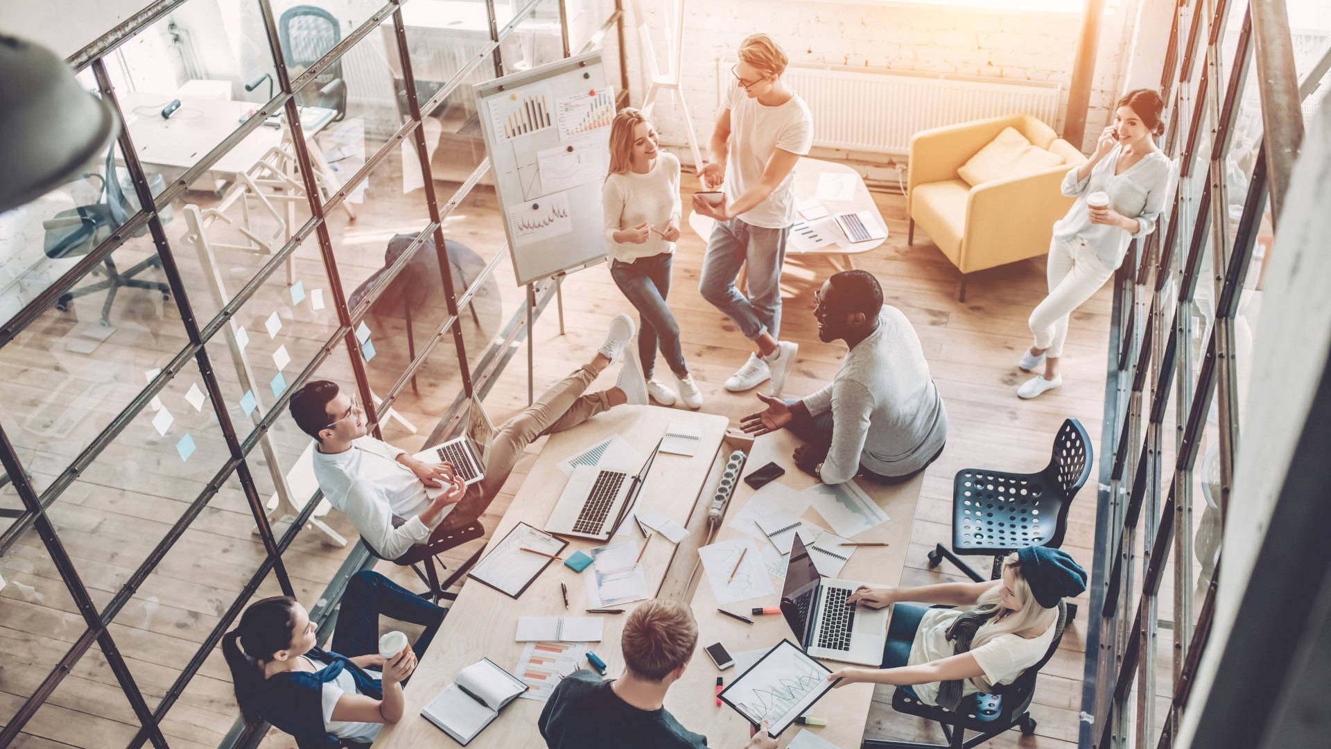 Ein modernes, lichtdurchflutetes Büro mit Glaswänden, in dem eine Gruppe junger Menschen an einem großen Tisch zusammenarbeitet. Laptops, Notizbücher und Diagramme liegen verteilt, während einige diskutieren, Ideen skizzieren oder auf einem Whiteboard präsentieren. Die lockere Atmosphäre und kreative Dynamik spiegeln einen Co-Creation- oder Design Thinking Workshop wider, bei dem kollaborativ innovative Lösungen entwickelt werden.