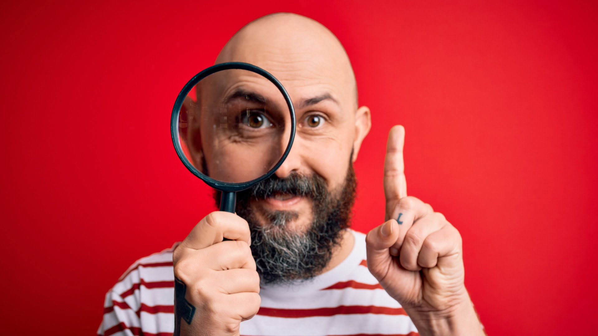 Bärtiger Mann mit Glatze in gestreiftem Shirt hält eine Lupe vor sein Auge und hebt mahnend den Finger vor rotem Hintergrund als Symbol für eine gute Strategie, die mit einer klaren Diagnose beginnt.