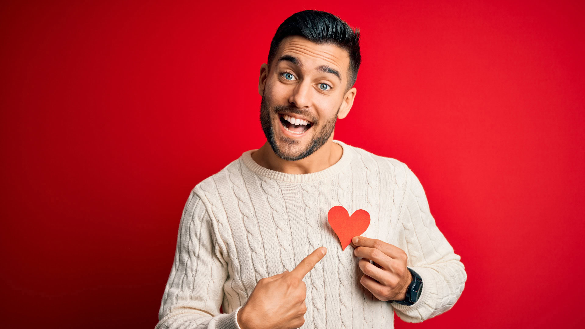 Ein lächelnder Mann mit kurzen dunklen Haaren und Bart, gekleidet in einen hellen Strickpullover, steht vor einem roten Hintergrund. Er hält ein rotes Papierherz an seine Brust und zeigt mit dem Finger darauf. Das Bild symbolisiert die Empathie im Design Thinking Prozess, bei dem Personas helfen, sich in die Bedürfnisse und Emotionen der Zielgruppe hineinzuversetzen.