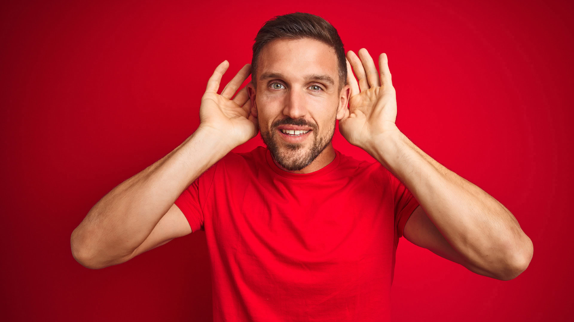Ein Mann in einem roten T-Shirt hält die Hände hinter seine Ohren, als würde er aufmerksam lauschen. Das Bild symbolisiert Social Listening, eine Technik zur Beobachtung von Online-Diskussionen, um die Bedürfnisse von Early Adopters besser zu verstehen.