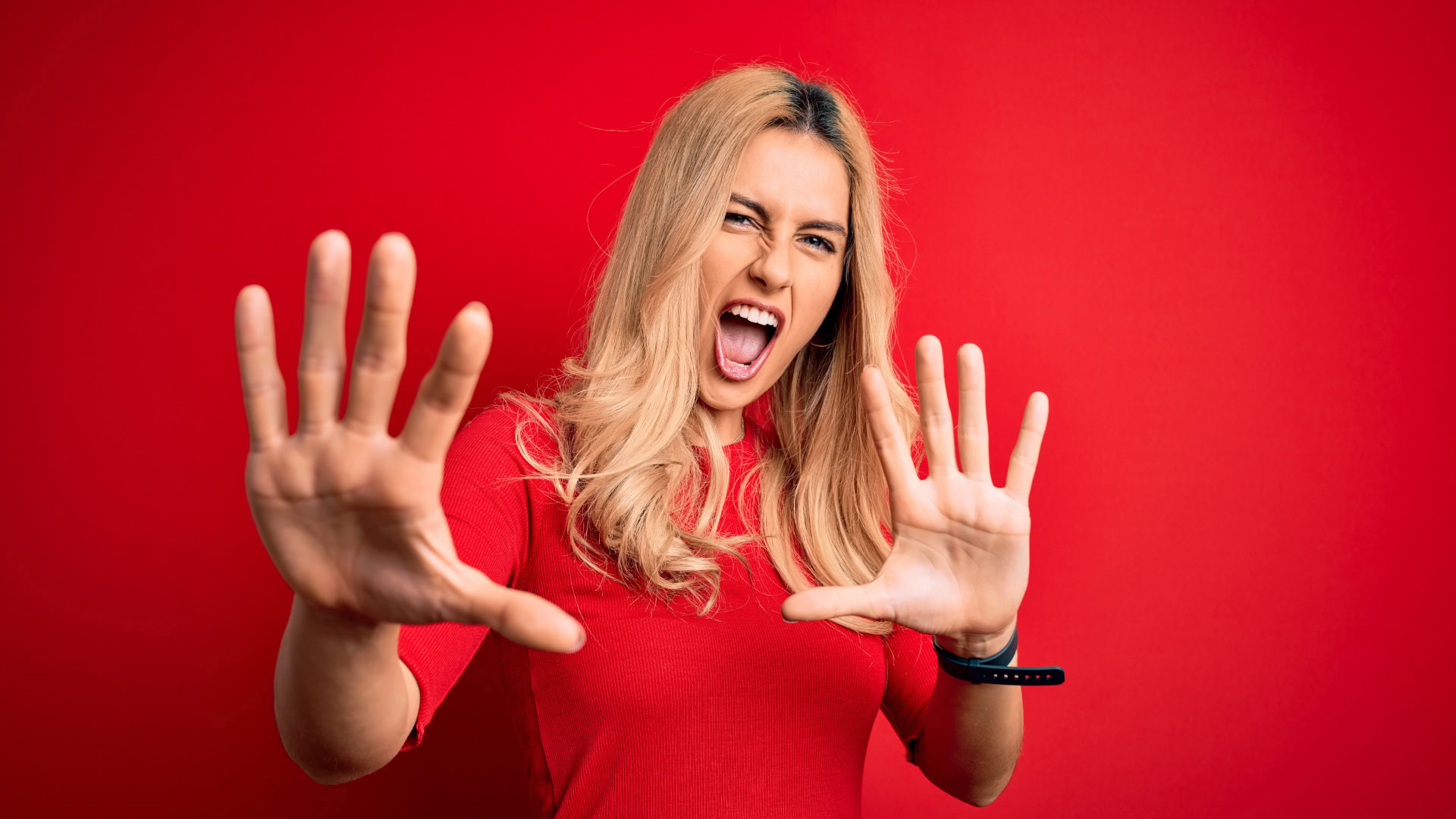 Eine Frau mit blondem Haar und rotem Oberteil steht vor einem roten Hintergrund und hebt beide Hände mit gespreizten Fingern abwehrend nach vorne, während sie mit ausdrucksstarkem Gesichtsausdruck den Mund öffnet. Das Bild symbolisiert ein Fixed Mindset und die Ablehnung von Herausforderungen, indem es zeigt, wie eine Person sich gegen neue oder ungewohnte Situationen sträubt.