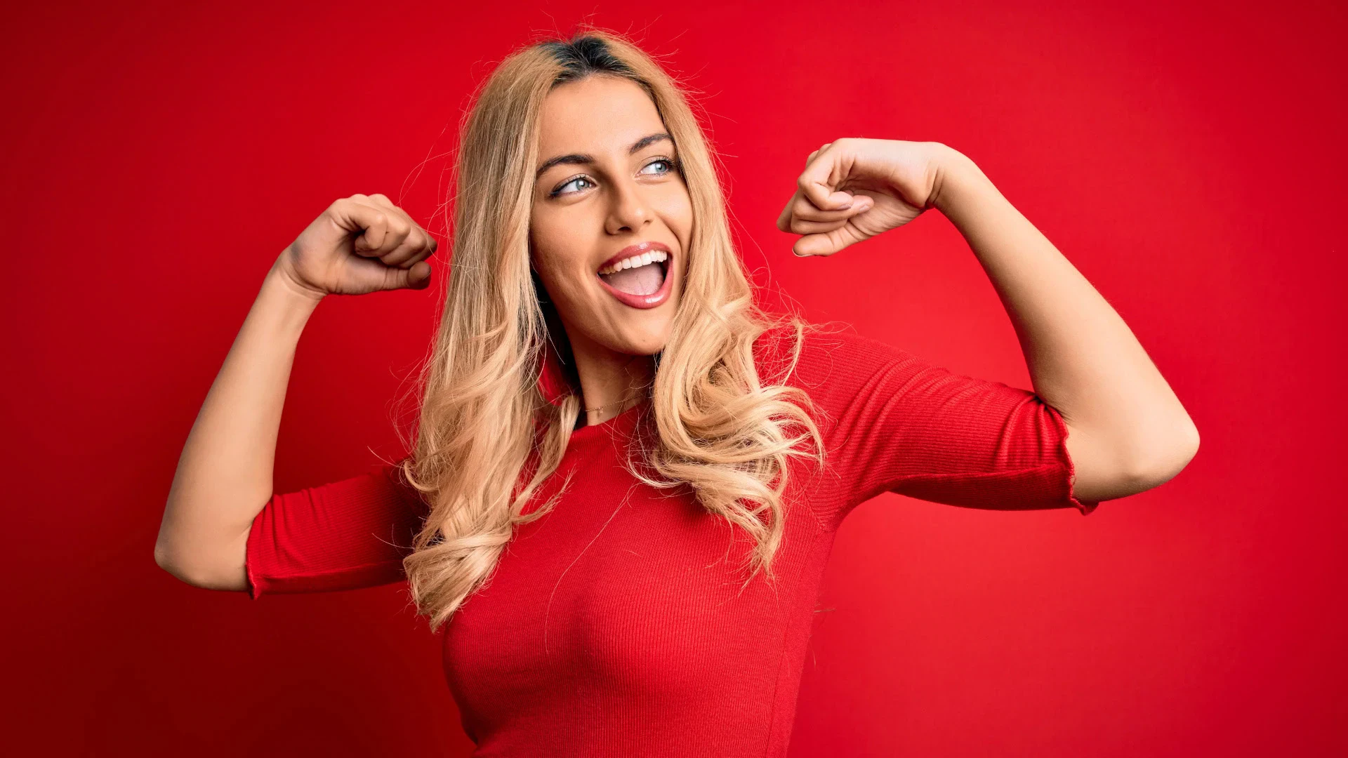 Eine selbstbewusste, lächelnde Frau in einem roten Oberteil steht vor einem roten Hintergrund und zeigt eine stolze, kraftvolle Pose mit angewinkelten Armen und geballten Fäusten. Das Bild symbolisiert ein Growth Mindset und Resilienz, indem es innere Stärke, positive Einstellung und die Fähigkeit zur persönlichen Weiterentwicklung visuell darstellt.