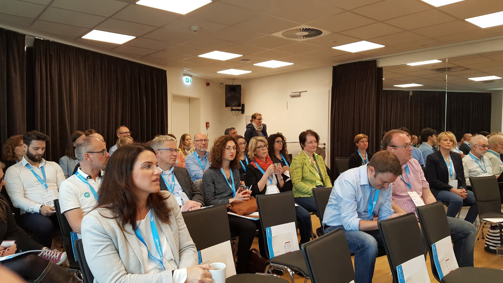 Eine Gruppe von Menschen sitzt in einem Konferenzraum mit schwarzen Vorhängen und heller Beleuchtung. Die Teilnehmer tragen Namensschilder an blauen Bändern und hören einer Präsentation zu. Einige machen sich Notizen oder halten Getränke in den Händen. Das Bild vermittelt die Atmosphäre eines Barcamps, bei dem sich die Teilnehmenden in einer offenen und interaktiven Umgebung austauschen.