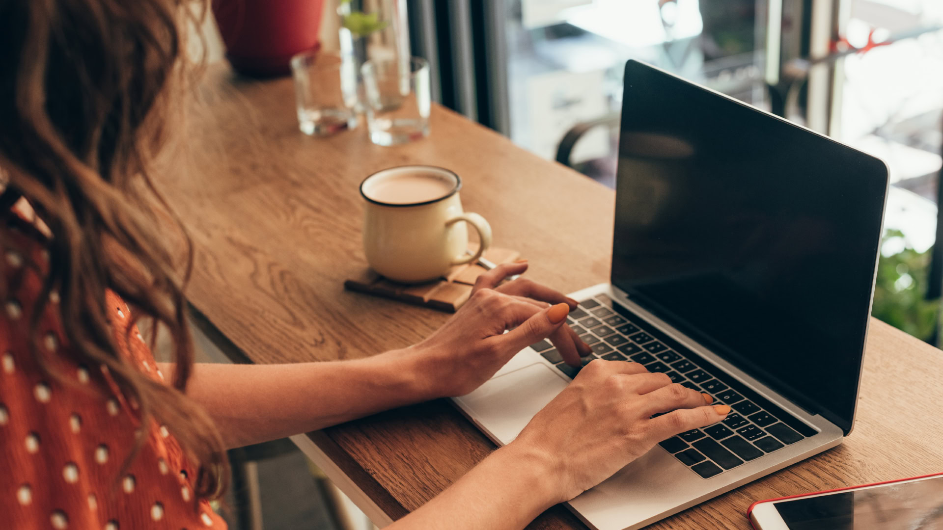 Eine junge rothaarige Frau sitzt in einem Café oder Co-Working-Space an einem Laptop und tippt konzentriert auf der Tastatur, während neben ihr eine Tasse Kaffee steht. Die Szene vermittelt den Kontext einer Job Story, indem sie eine typische Arbeitssituation zeigt, in der eine Aufgabe oder ein Ziel verfolgt wird.