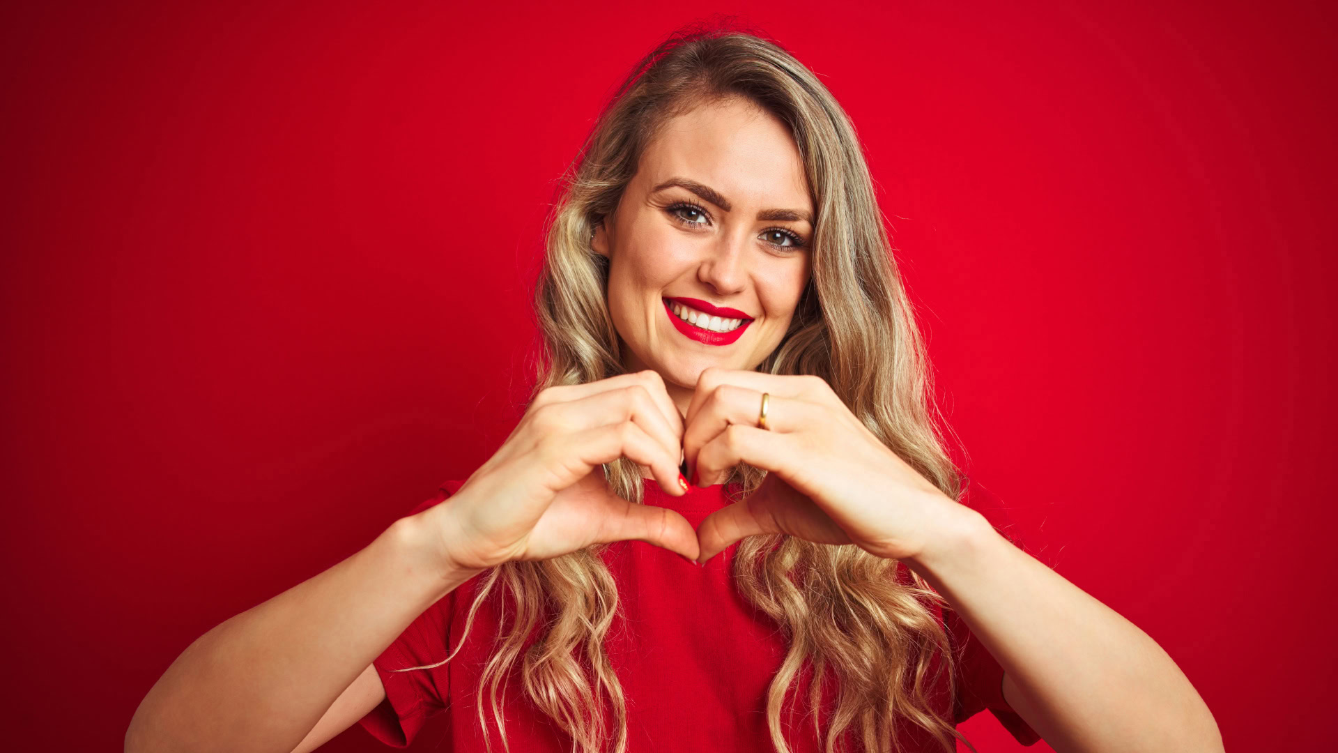 Eine lächelnde Frau in roter Kleidung vor einem roten Hintergrund formt mit ihren Händen ein Herzsymbol. Das Bild steht symbolisch für erfüllte Kundenwünsche und die emotionale Bindung zwischen einem Unternehmen und seinen Kunden. Zufriedene Kunden fühlen sich wertgeschätzt und entwickeln eine stärkere Loyalität zur Marke.