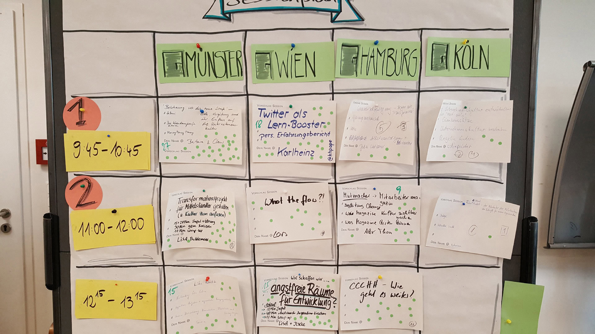 Ein Session-Plan auf einem Whiteboard zeigt die Agenda eines Barcamps. Die einzelnen Zeit-Slots sind auf gelben Karten notiert, während darunter handgeschriebene Kärtchen mit Session-Vorschlägen und Namen der Moderatoren angepinnt sind.