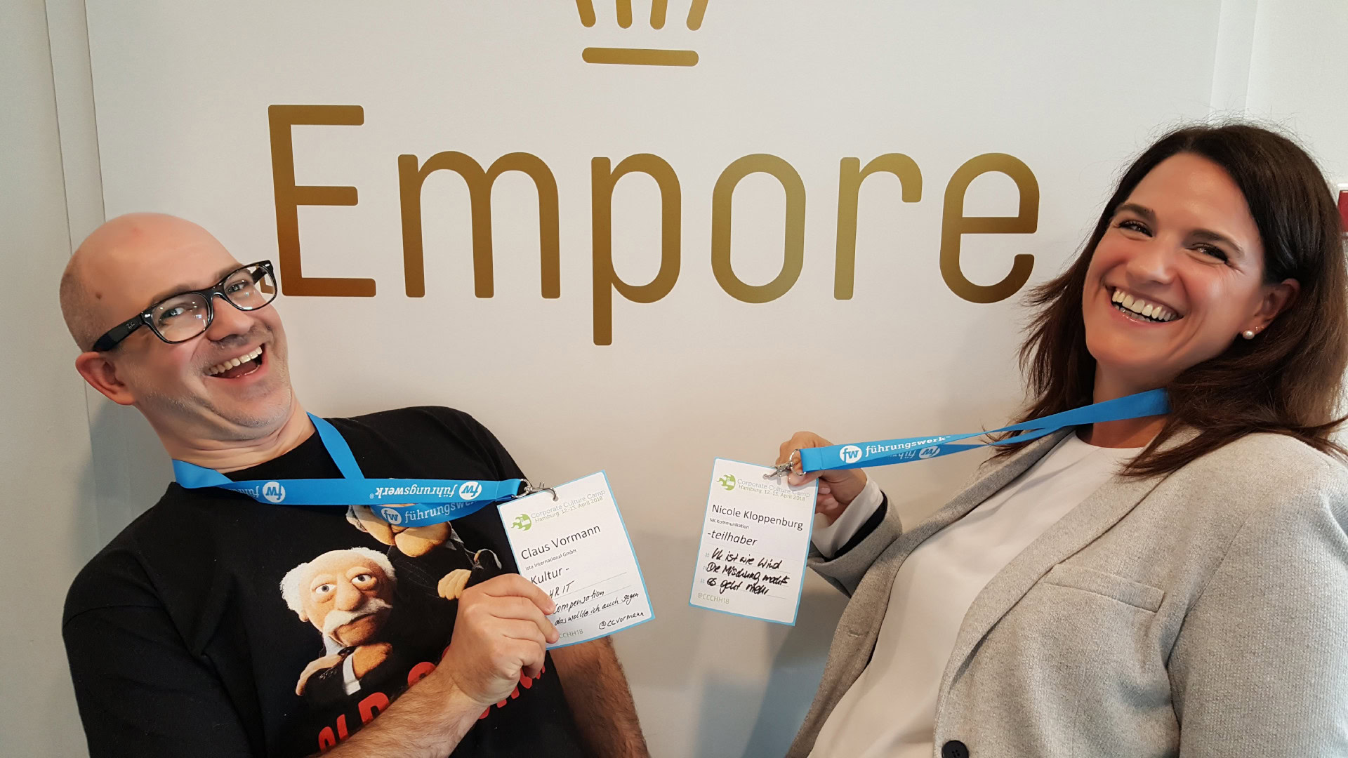 Zwei lachende Personen mit blauen Namensschildern stehen vor einer weißen Wand mit goldener Schrift. Sie zeigen ihre Teilnehmerausweise eines Barcamps und strahlen Begeisterung aus. Die beiden Personen spielen das Synergie-Partnerspiel, bei dem Teilnehmende passende Gesprächspartner für den Austausch und die Zusammenarbeit finden.