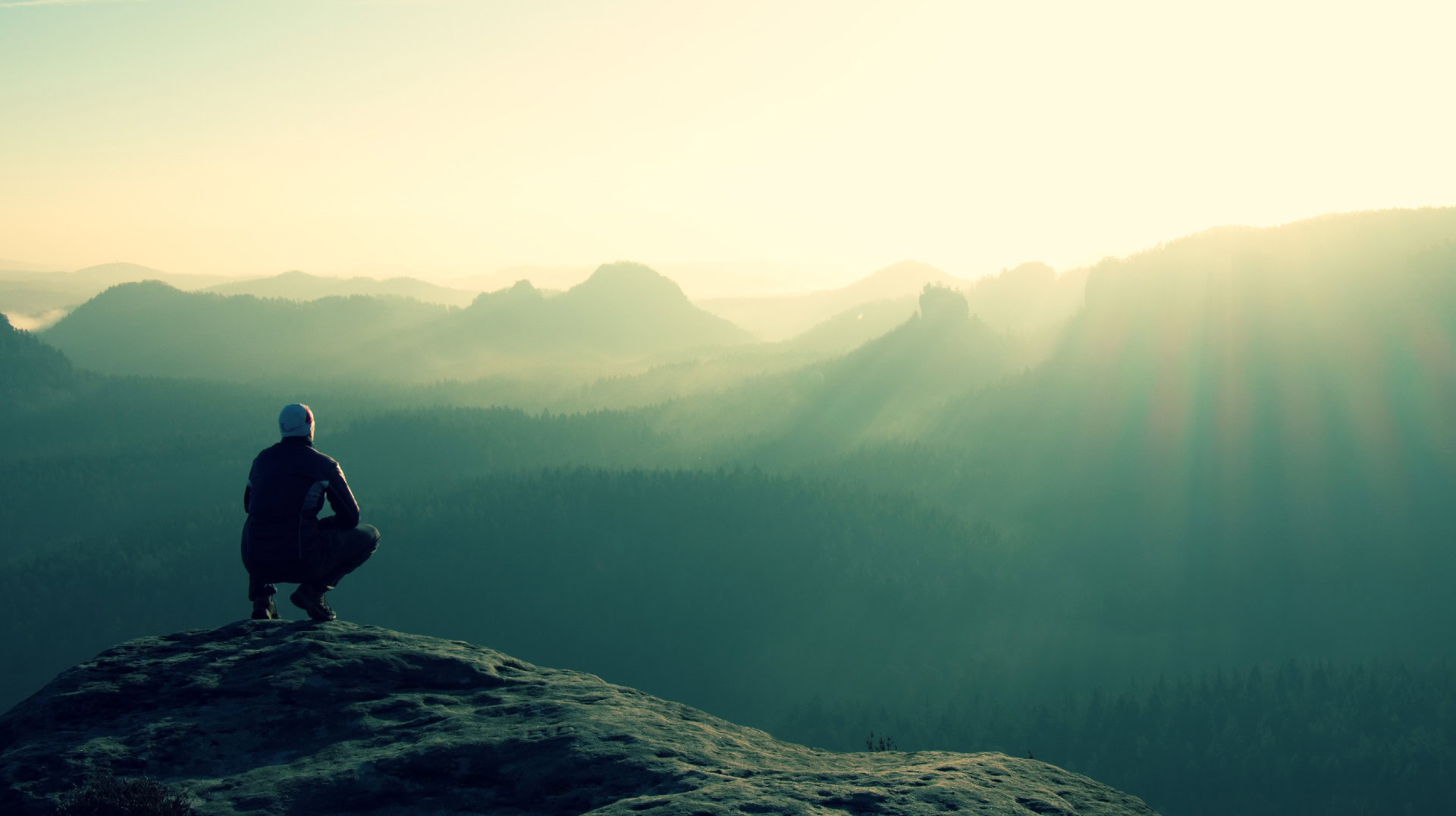 Ein Mensch hockt auf einem Felsen und blickt über eine weite, neblige Berglandschaft, während die Sonnenstrahlen den Horizont erhellen. Das Bild symbolisiert, wie wichtig es ist, Ziele zu formulieren, die nicht nur erreichbar sind, sondern nach ihrer Verwirklichung neue Perspektiven eröffnen und den Blick auf bisher unbekannte Möglichkeiten freigeben.