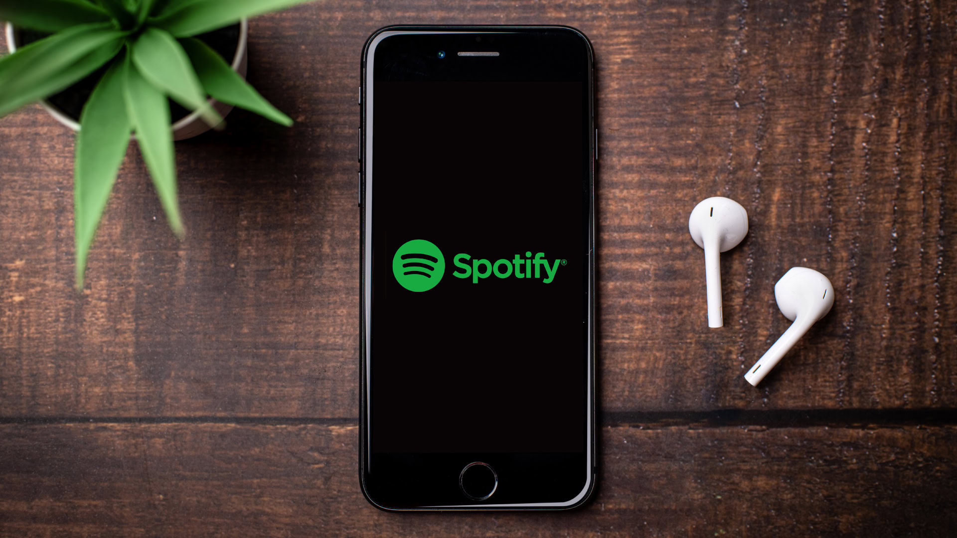 Ein Handy liegt auf einem Holztisch, daneben zwei weiße In-Ear-Kopfhörer. Auf dem Display ist das Spotify-Logo zu sehen.