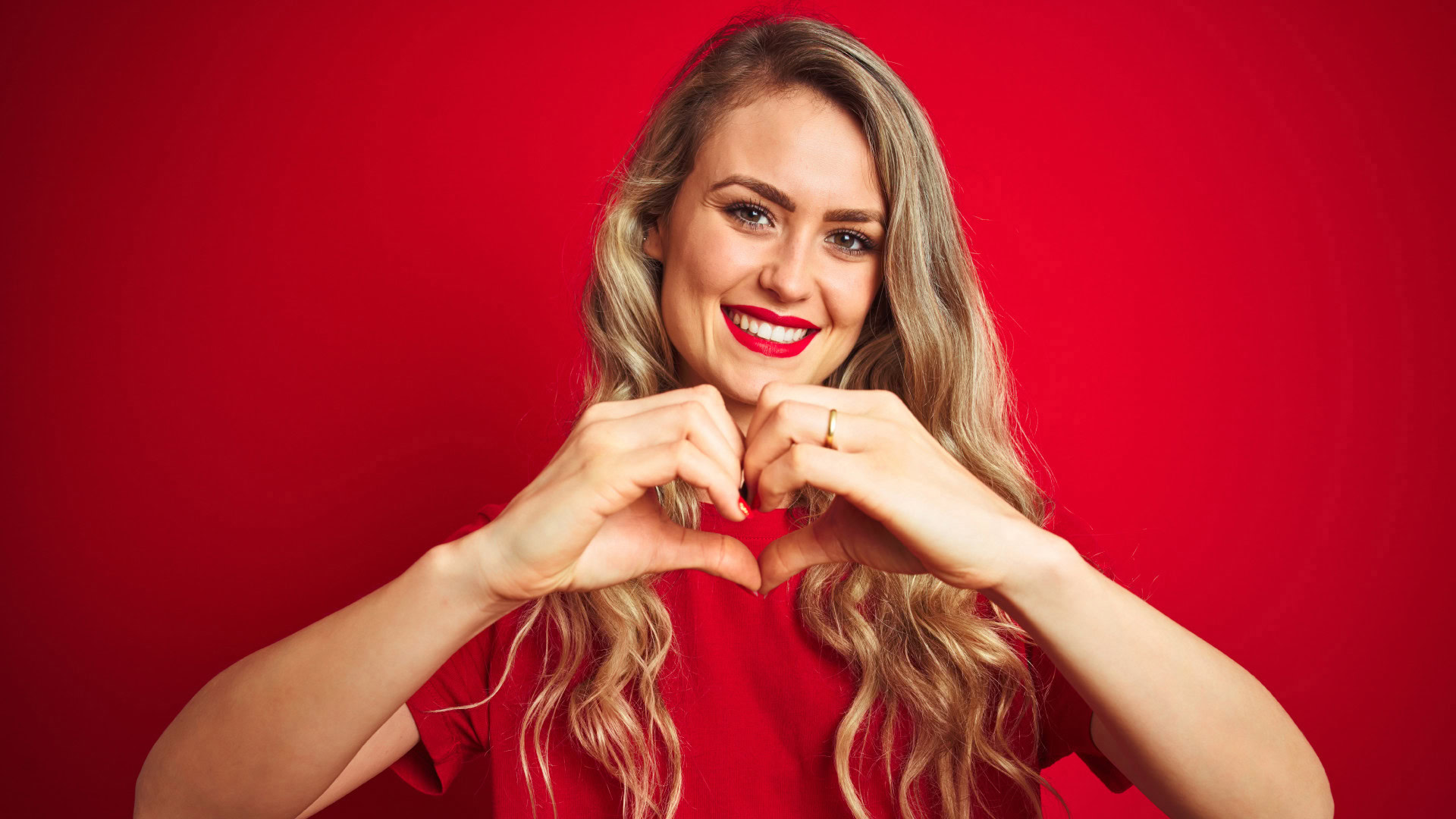 Strahlende Frau mit langen, gewellten blonden Haaren und rotem Lippenstift formt mit ihren Händen ein Herz vor einem roten Hintergrund, symbolisiert die Kraft gut formulierter OKR, die Resonanz und Inspiration erzeugen.