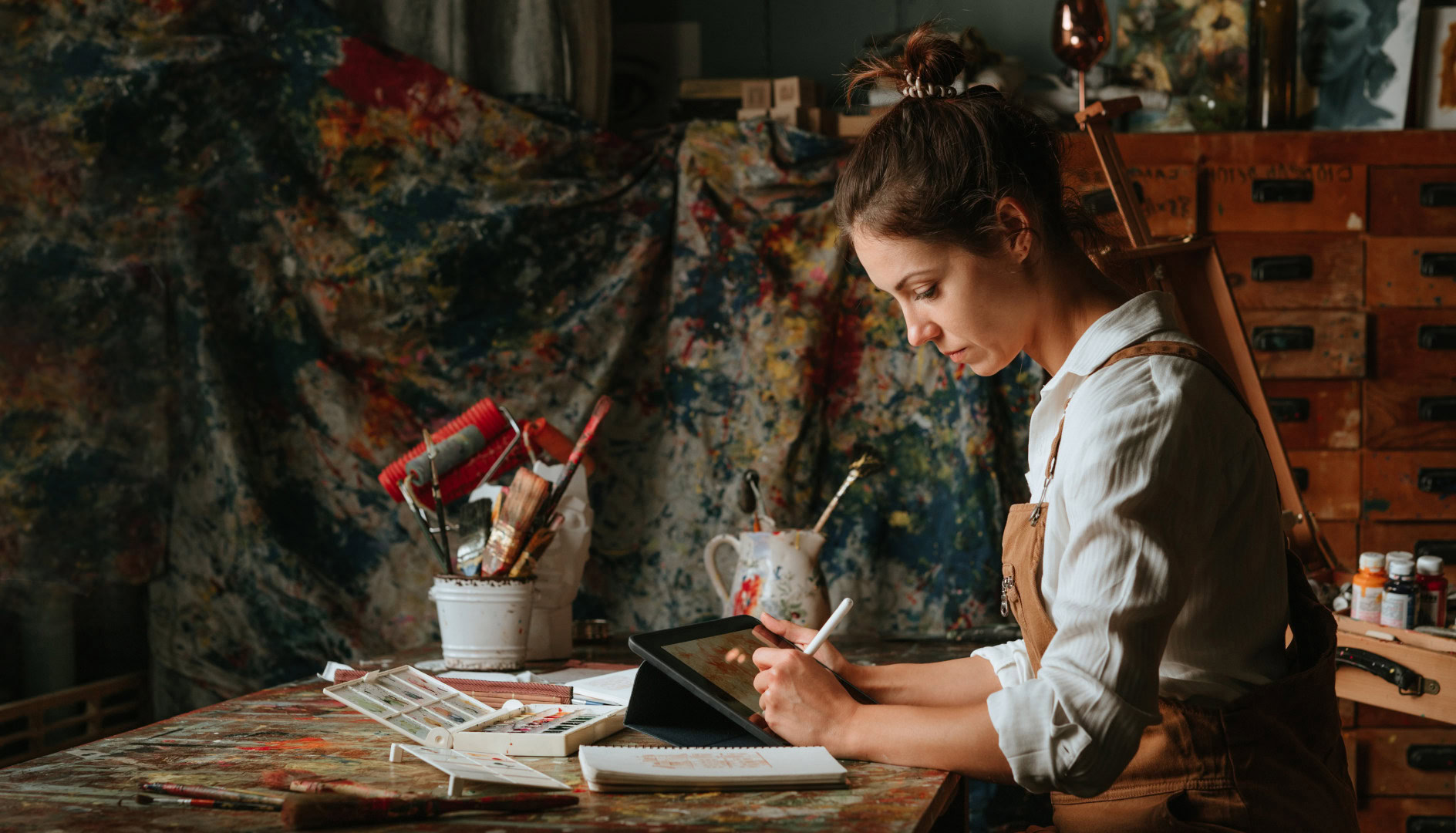 Eine Hobby-Künstlerin sitzt in einem kreativen Atelier und arbeitet konzentriert mit einem digitalen Zeichenstift auf einem Tablet. Um sie herum liegen Pinsel, Farben und Zeichenutensilien, während der Hintergrund mit Farbspritzern bedeckt ist.