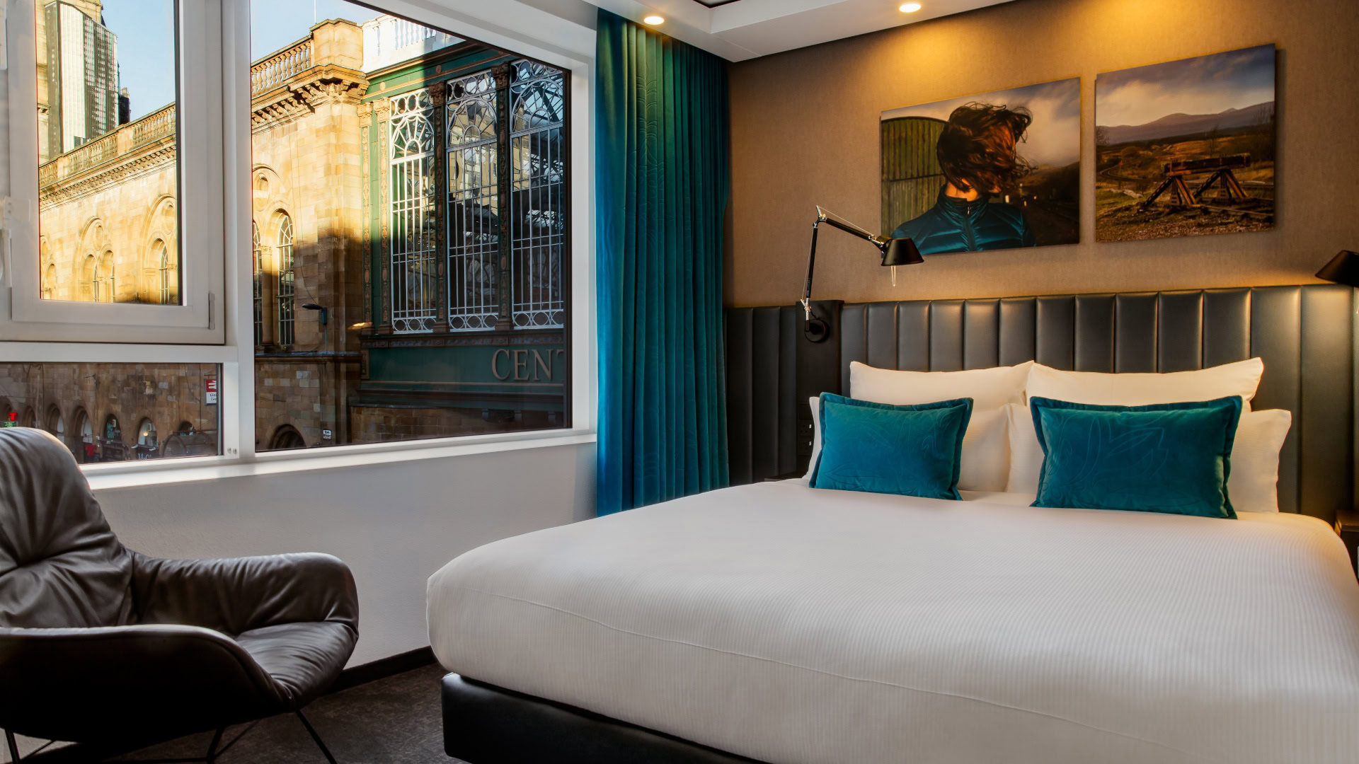 Ein stilvolles Hotelzimmer von Motel One in Glasgow mit modernem Design. Im Mittelpunkt steht ein großes Bett mit weißen Bezügen und blauen Kissen, das von einer gepolsterten Wand und stimmungsvoller Beleuchtung eingerahmt wird. Durch das Fenster fällt der Blick auf eine historische Fassade. Das Bild spiegelt das innovative Geschäftsmodell von Motel One wider, das Design, Komfort und erschwingliche Preise kombiniert.