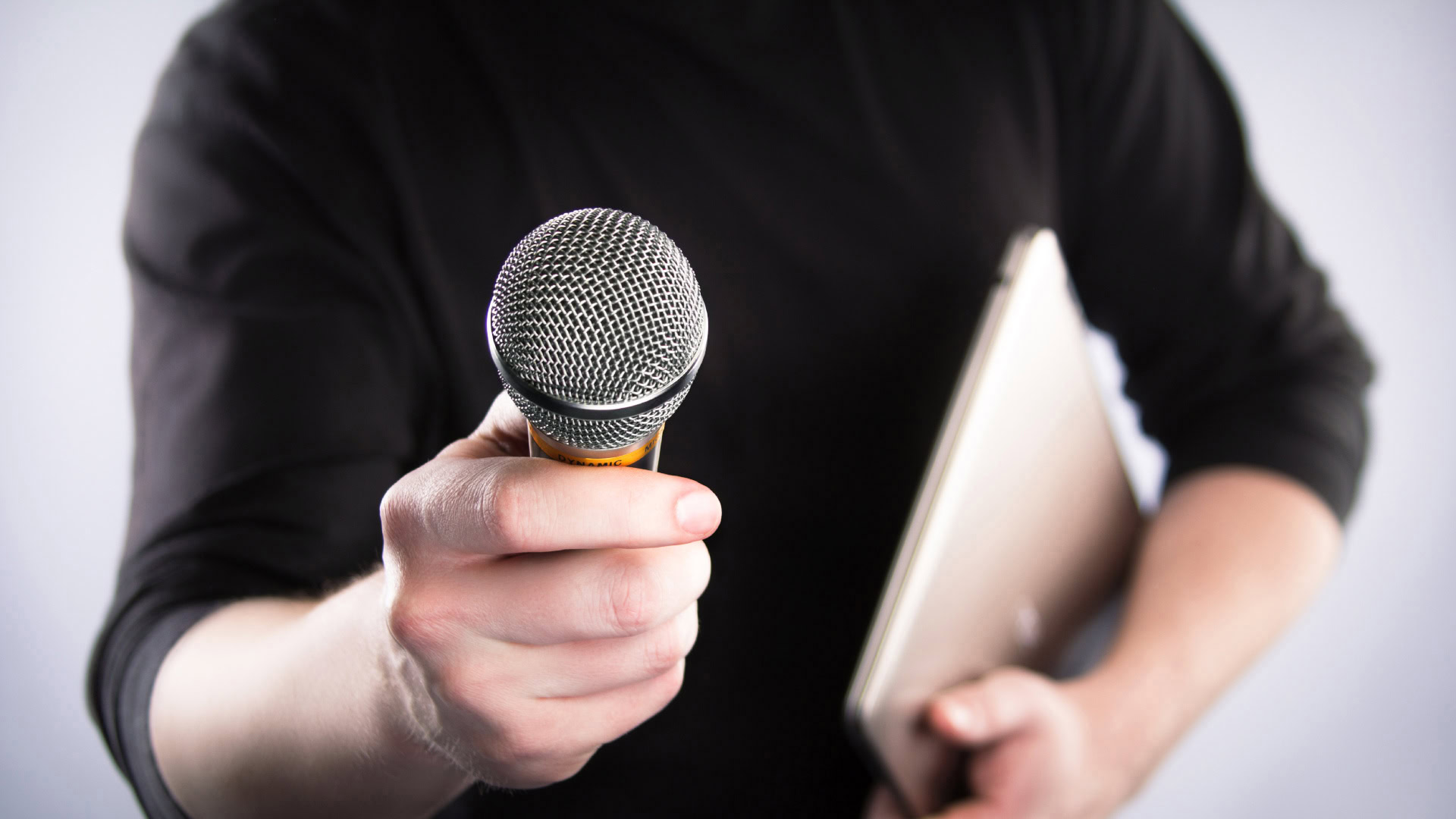 Eine Person in schwarzer Kleidung hält ein Mikrofon direkt in die Kamera und trägt in der anderen Hand ein Tablet oder Notizbuch. Der Fokus liegt auf dem Mikrofon, während der Hintergrund und die Person leicht unscharf sind.