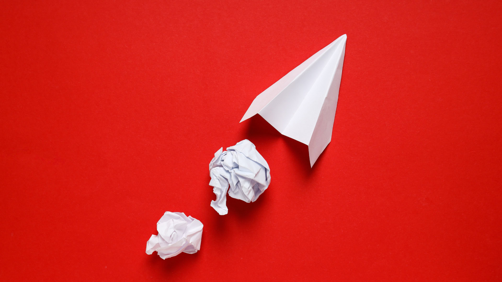 Ein weißer Papierflieger auf rotem Hintergrund mit zwei zerknüllten Papierbällen davor, symbolisiert iteratives Vorgehen und den Lernprozess durch Versuch und Irrtum.