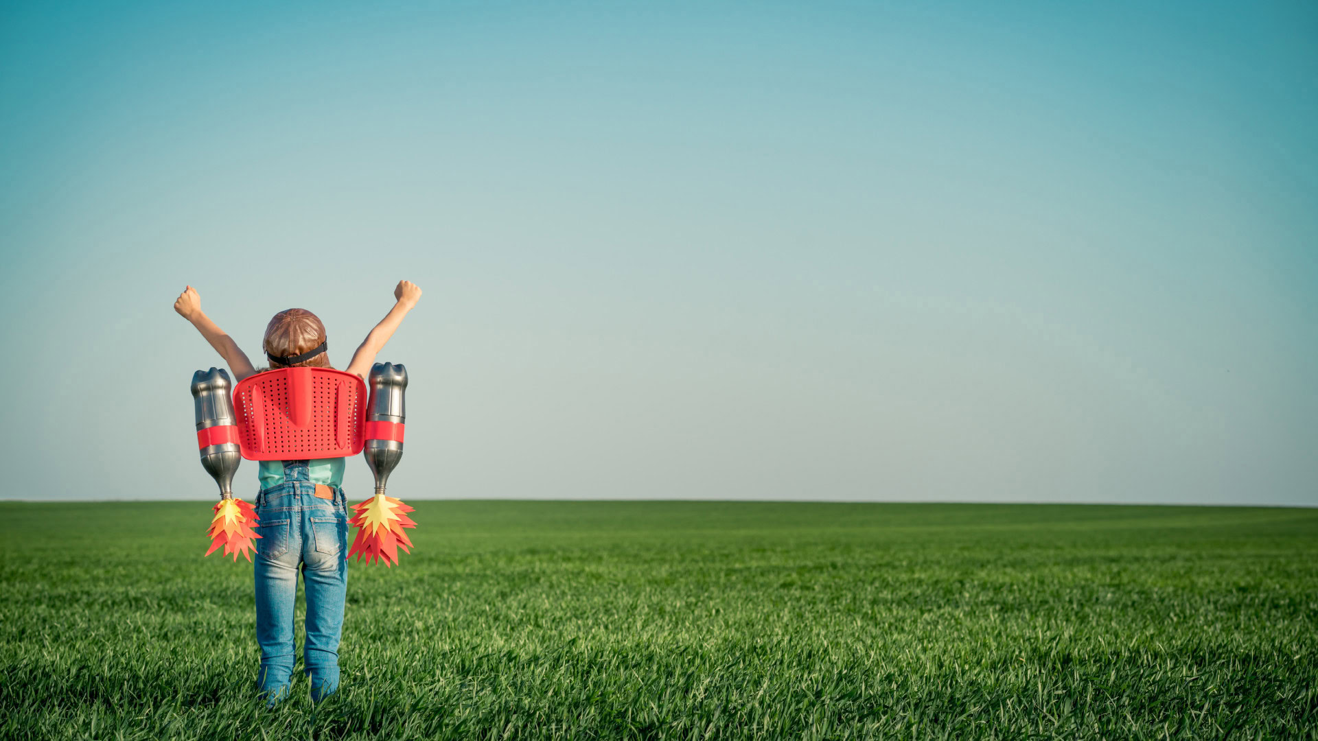 Ein Kind mit ausgestreckten Armen trägt einen selbstgebastelten Raketenrucksack aus Alltagsgegenständen und steht auf einer weiten grünen Wiese unter einem klaren Himmel, symbolisiert die unbeschränkte Kreativität und divergentes Denken.