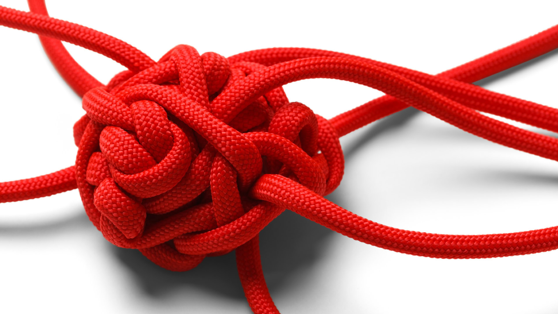 Ein dicht verknotetes rotes Seil mit mehreren losen Enden liegt auf einem weißen Hintergrund. Der Knoten wirkt unübersichtlich und schwer entwirrbar. Dieses Bild steht symbolisch für Komplexität, da die vielen miteinander verknüpften Elemente ein unvorhersehbares und dynamisches System darstellen, das nicht einfach mit einer festen Anleitung gelöst werden kann.