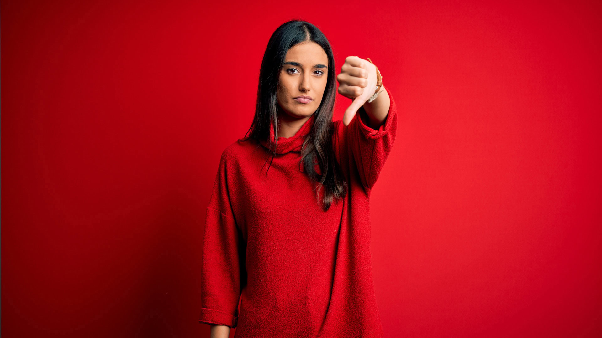 Eine Frau mit langen dunklen Haaren, die einen roten Pullover trägt und vor einem roten Hintergrund steht. Sie zeigt mit neutralem Gesichtsausdruck einen Daumen nach unten. Dieses Bild symbolisiert, dass nicht alle Kundensegmente gleichermaßen begeistert sind und es wichtig ist, die Bedürfnisse und Erwartungen verschiedener Zielgruppen zu verstehen.