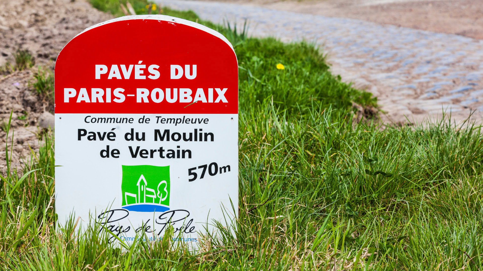 OKR Beispiel mit einer Metapher für schlechte Key Results. Ein Schild am Streckenrand des berühmten Radrennens Paris-Roubaix markiert einen 570 Meter langen Kopfsteinpflasterabschnitt. Symbolisiert das Problem von aufeinander aufbauenden Key Results, die wie Meilensteine nacheinander erreicht werden müssen, anstatt unabhängig messbare Fortschritte darzustellen.