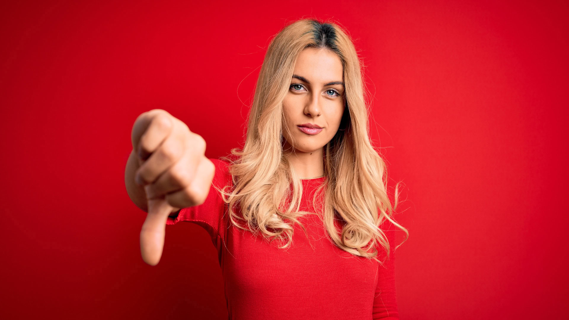 Frau mit blondem Haar und rotem Pullover zeigt den Daumen nach unten vor rotem Hintergrund als Symbol für eine gute Strategie, klare Priorisierung und den Fokus auf das Wesentliche durch bewusstes Nein-Sagen