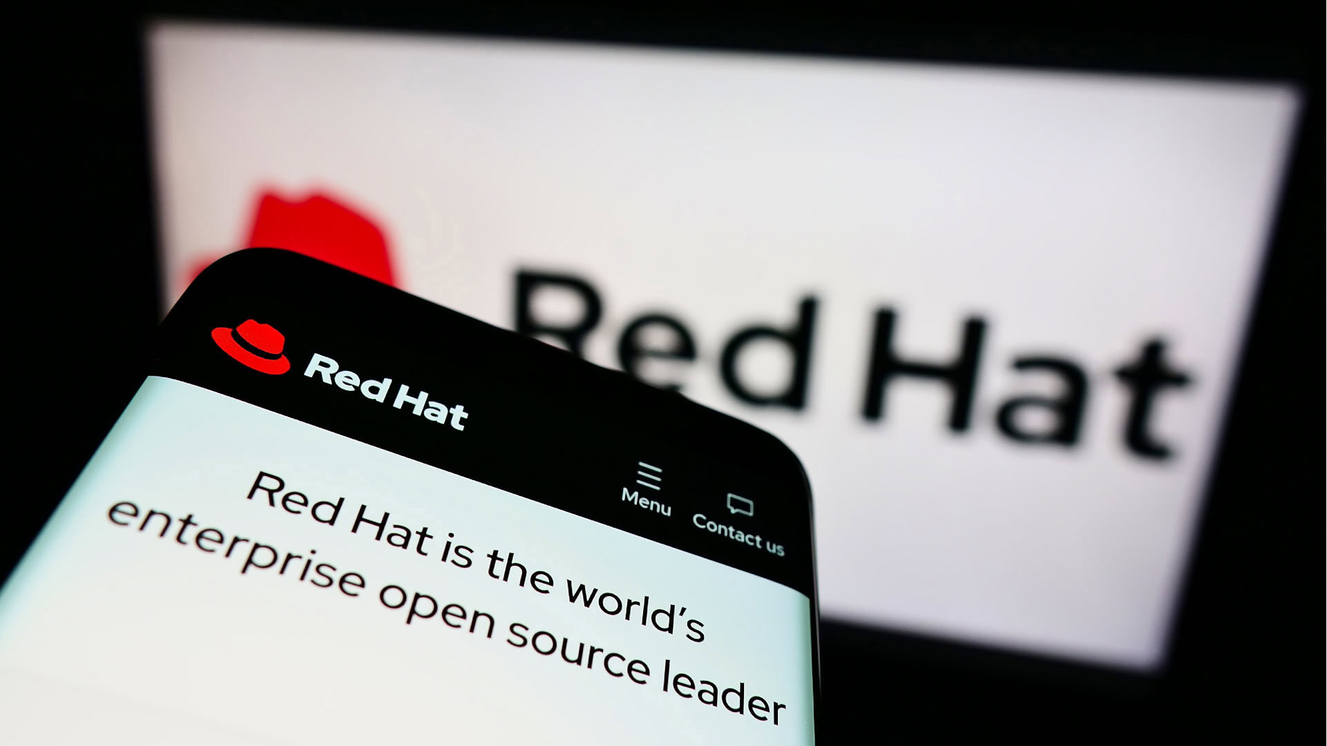 Red Hat als Beispiel für das Open-Source-Geschäftsmodell. Im Vordergrund ist ein Smartphone zu sehen, das die Website von Red Hat mit dem Slogan „Red Hat is the world's enterprise open source leader“ anzeigt. Im Hintergrund erscheint das Red-Hat-Logo verschwommen auf einem größeren Bildschirm. Red Hat nutzt das Open-Source-Geschäftsmodell, indem es freie Software bereitstellt und durch zusätzliche Dienstleistungen wie Support, Beratung und maßgeschneiderte Lösungen Umsatz generiert.