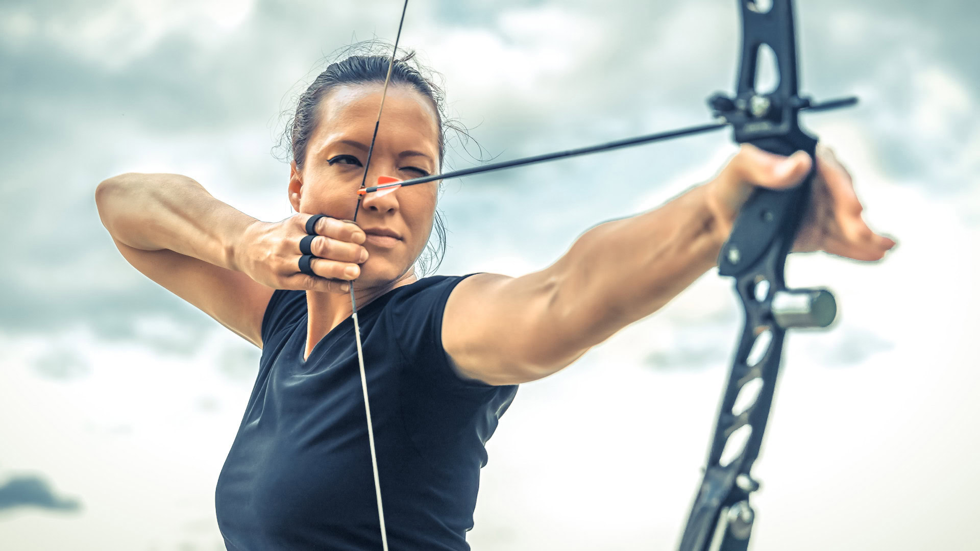 Eine fokussierte Frau mit dunklen Haaren und einem schwarzen Sportshirt spannt einen modernen Bogen und zielt konzentriert in die Ferne. Im Hintergrund sind Wolken am Himmel zu sehen. Das Bild symbolisiert, dass auch Hobbys und persönliche Interessen in einer Design Thinking Persona berücksichtigt werden können, um ein ganzheitliches Bild der Zielgruppe zu erhalten.