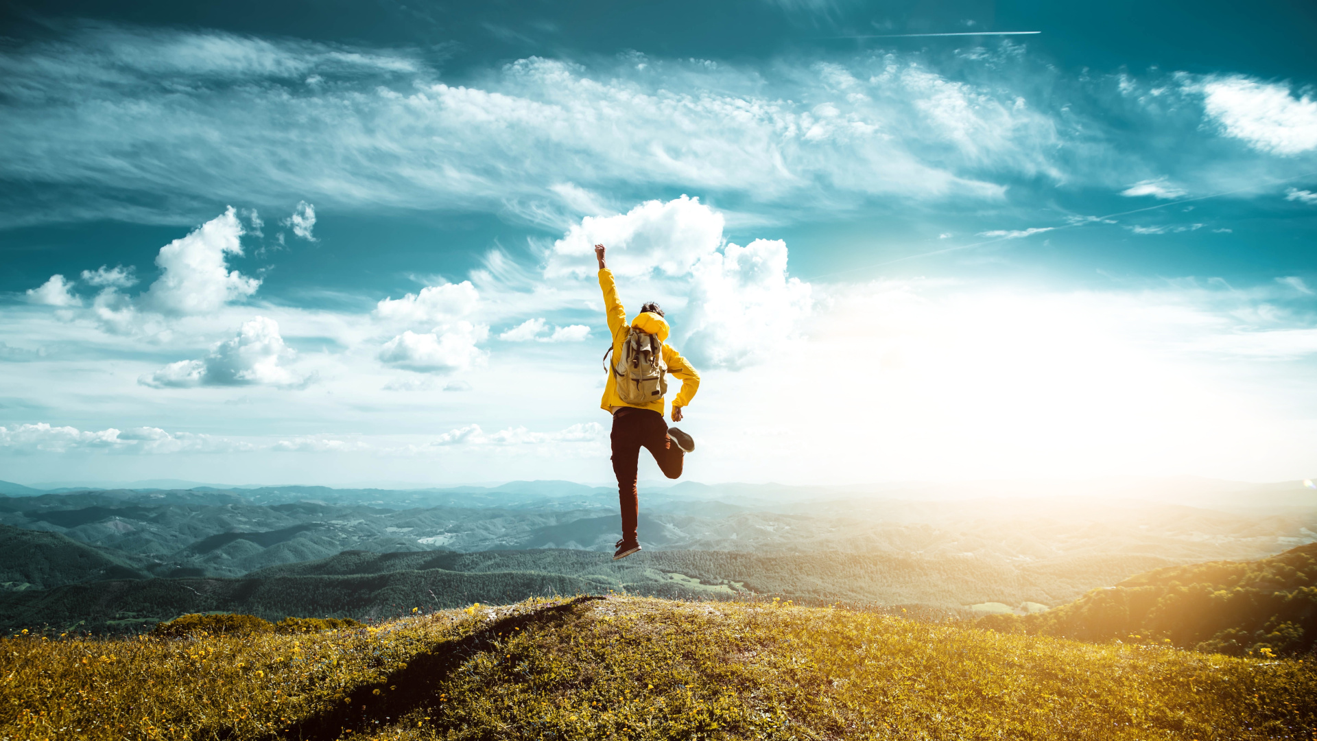 Ein Solopreneur in einer gelben Jacke und mit Rucksack springt euphorisch auf einem Berggipfel in die Luft. Im Hintergrund erstreckt sich eine weite Hügellandschaft unter einem strahlend blauen Himmel.