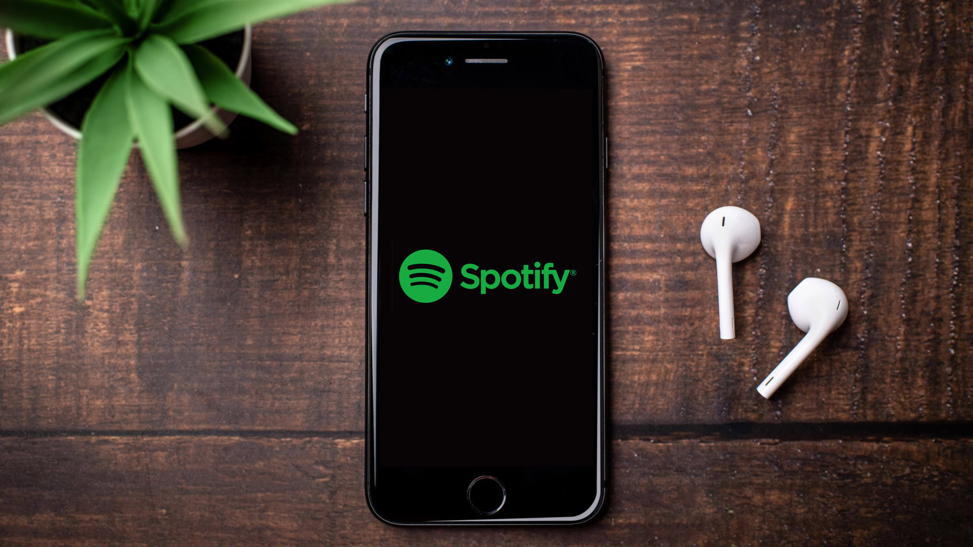 Ein Handy liegt auf einem Holztisch, daneben zwei weiße In-Ear-Kopfhörer. Auf dem Display ist das Spotify-Logo zu sehen.
