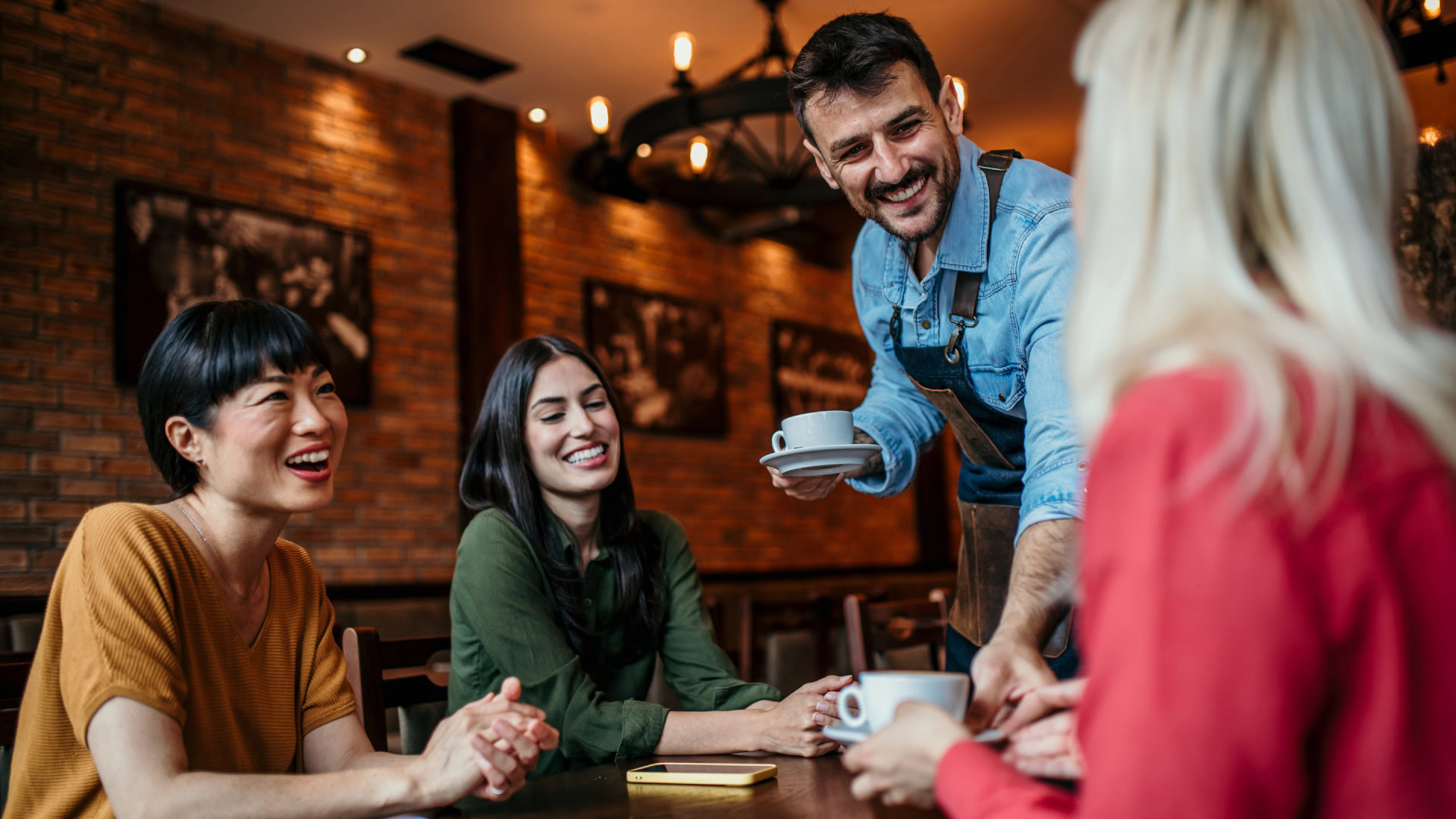 Ein Barista serviert Kaffee an eine Gruppe fröhlicher Menschen in einem gemütlichen Café, was die lockere und offene Atmosphäre eines User Interviews widerspiegelt.