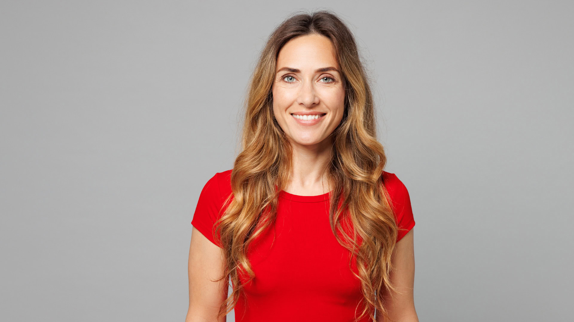 Porträt einer lächelnden Frau mit langen, welligen Haaren in einem roten Shirt vor neutralem grauem Hintergrund. Dieses Stockfoto dient als visuelle Darstellung für die User Persona Rebecca Holtkamp, eine fiktive Zielgruppen-Vertreterin.