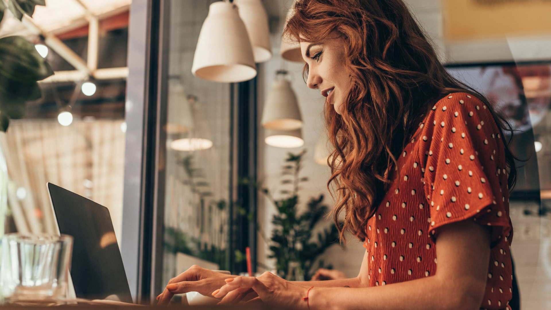 Eine junge Frau mit langen, lockigen roten Haaren arbeitet konzentriert an einem Laptop in einem stilvoll eingerichteten Café. Die Umgebung ist modern mit Hängelampen, Glasfront und Pflanzen. Dieses Bild symbolisiert die User Persona im realen Nutzungskontext, um ein besseres Verständnis für deren Arbeitsweise und Umgebung zu erhalten.