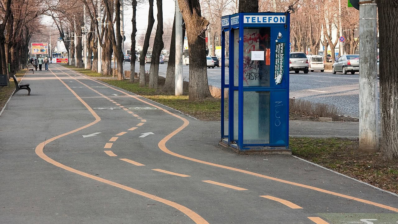 Ein Fahrradweg mit gelben Markierungen verläuft gerade, bis er plötzlich in Schlangenlinien um eine blaue, veraltete Telefonzelle herumführt, bevor er sich wieder begradigt. Die absurde Streckenführung zeigt einen Workaround, der eine ungeplante Lösung für ein Hindernis darstellt.