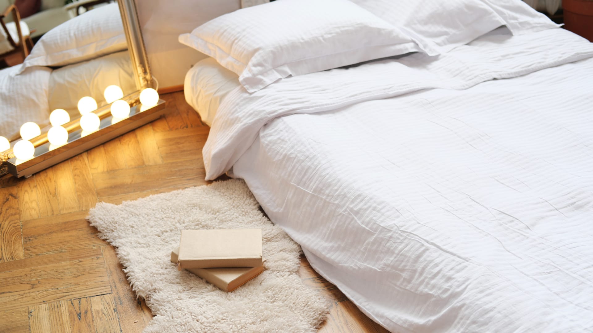 Gemütlich eingerichtetes Schlafstätte mit einer weißen Luftmatratze auf Holzboden, einem flauschigen Teppich, zwei geschlossenen Büchern und einem beleuchteten Spiegel im Hintergrund.