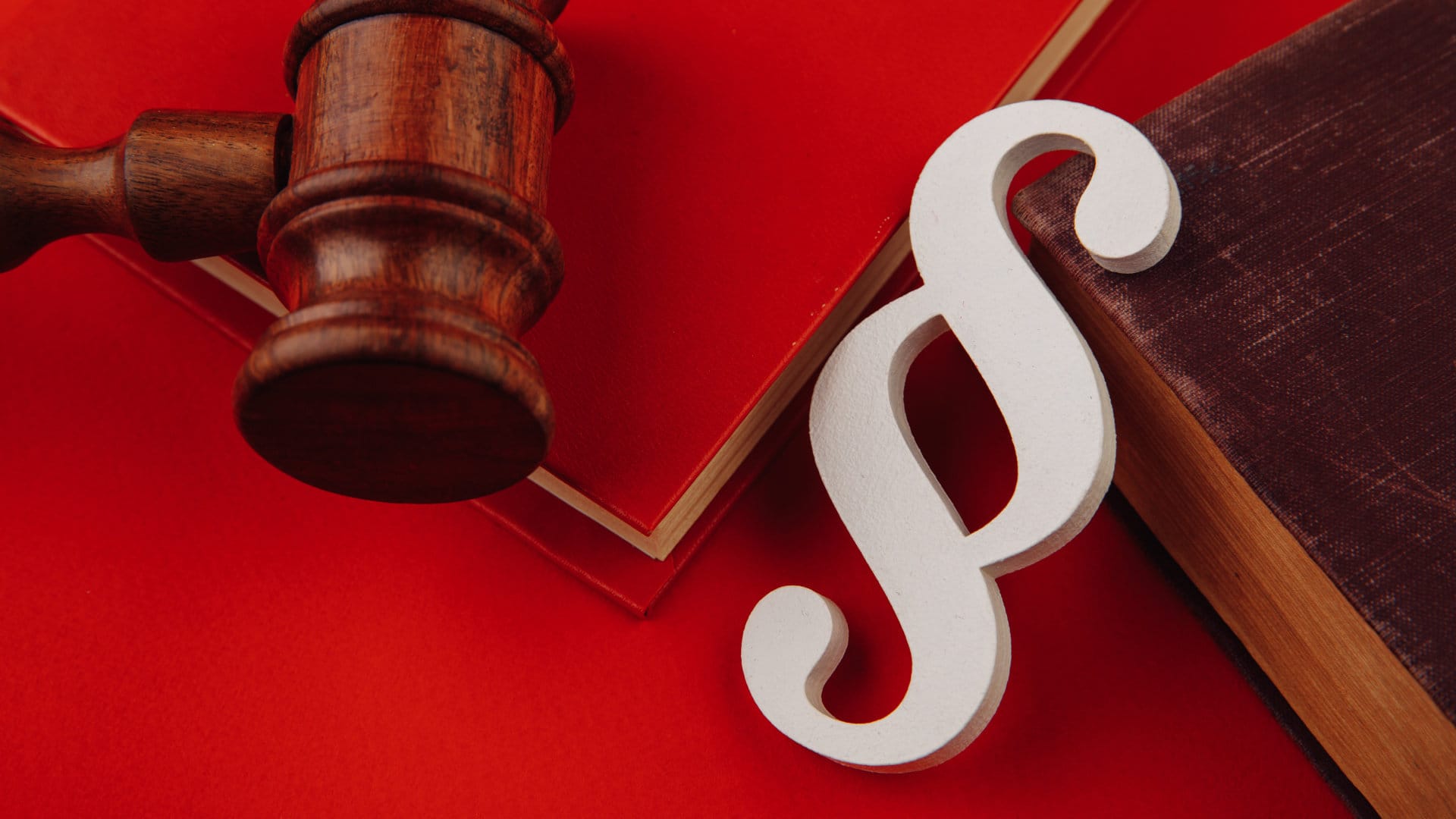 Holzhammer, Paragrafenzeichen und Gesetzesbücher auf rotem Hintergrund.