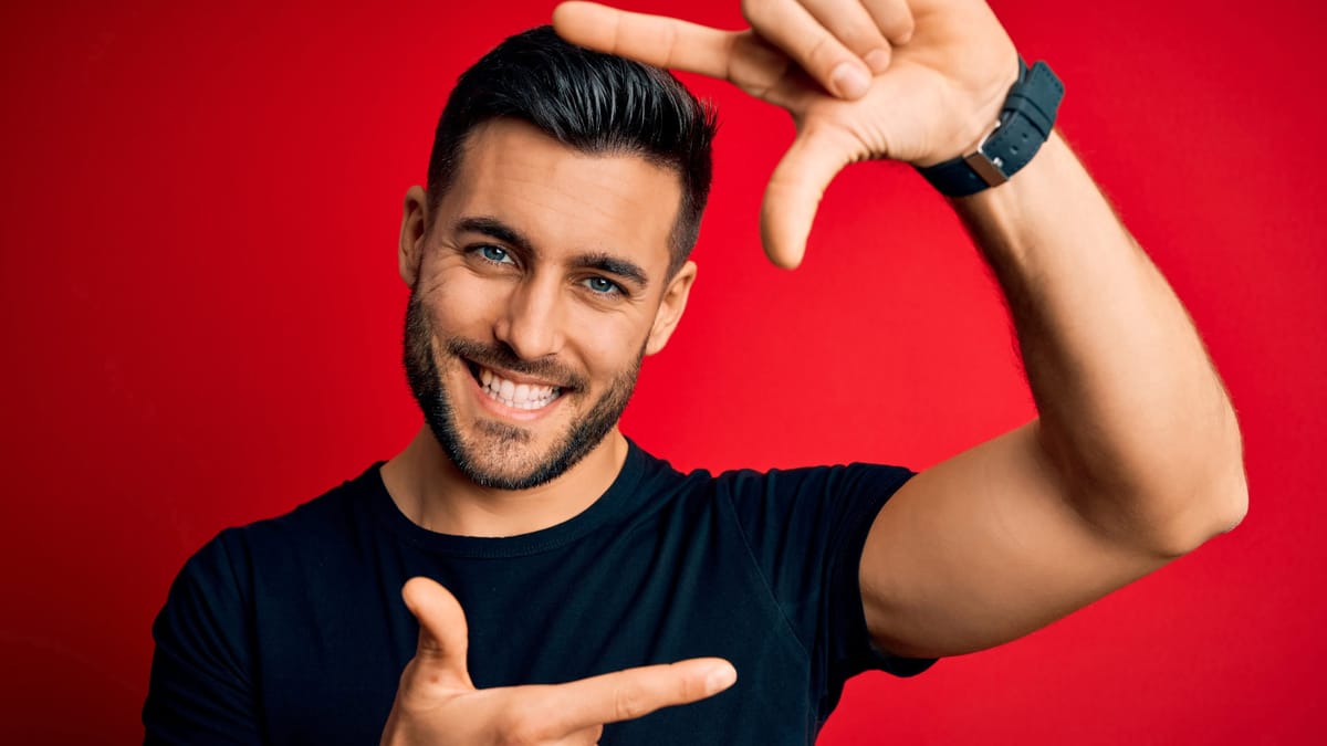 Ein lächelnder Mann in schwarzem Shirt vor rotem Hintergrund formt mit seinen Händen einen imaginären Rahmen, der das Konzept