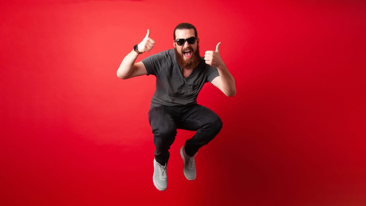 Ein Mann mit Bart, Sonnenbrille und dunkler Kleidung springt in die Luft und zeigt mit beiden Händen einen Daumen hoch vor ei