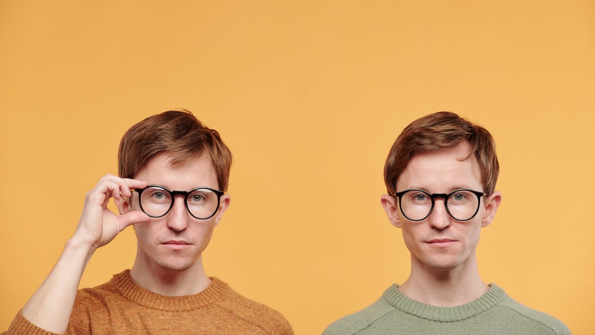 Zwei Zwillinge mit Brillen und ähnlicher Kleidung stehen vor einem orangefarbenen Hintergrund. Ihre fast identische Erscheinu