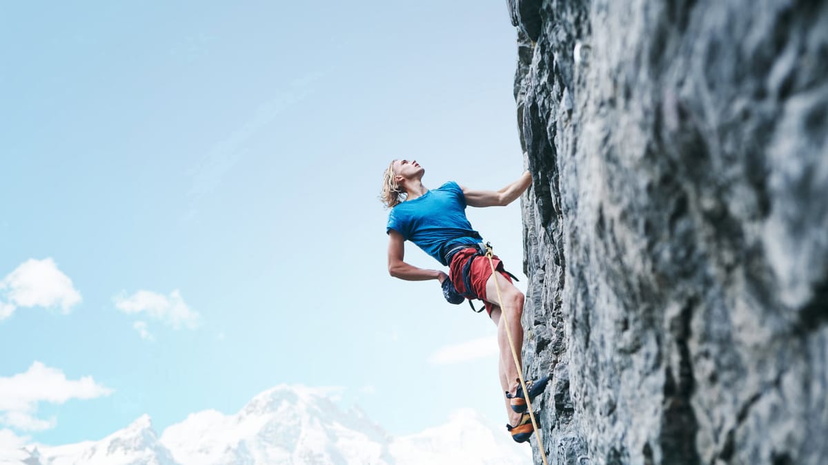 Ein Kletterer mit blonden Haaren in blauer Sportkleidung und roter Kletterhose erklimmt eine steile Felswand, während er nach