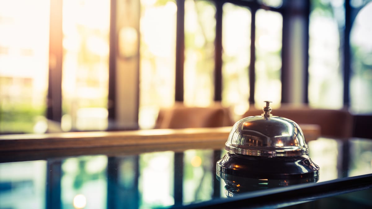 Silberne Hotelklingel steht auf einem glänzenden Tresen, im Hintergrund weiches Sonnenlicht und eine unscharfe Fensterfront.