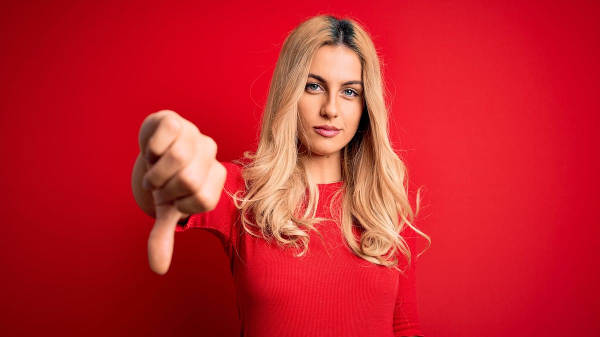 Junge Frau mit blondem Haar zeigt mit ernstem Blick den Daumen nach unten. Der rote Hintergrund und ihr rotes Oberteil betonen die ablehnende Geste.