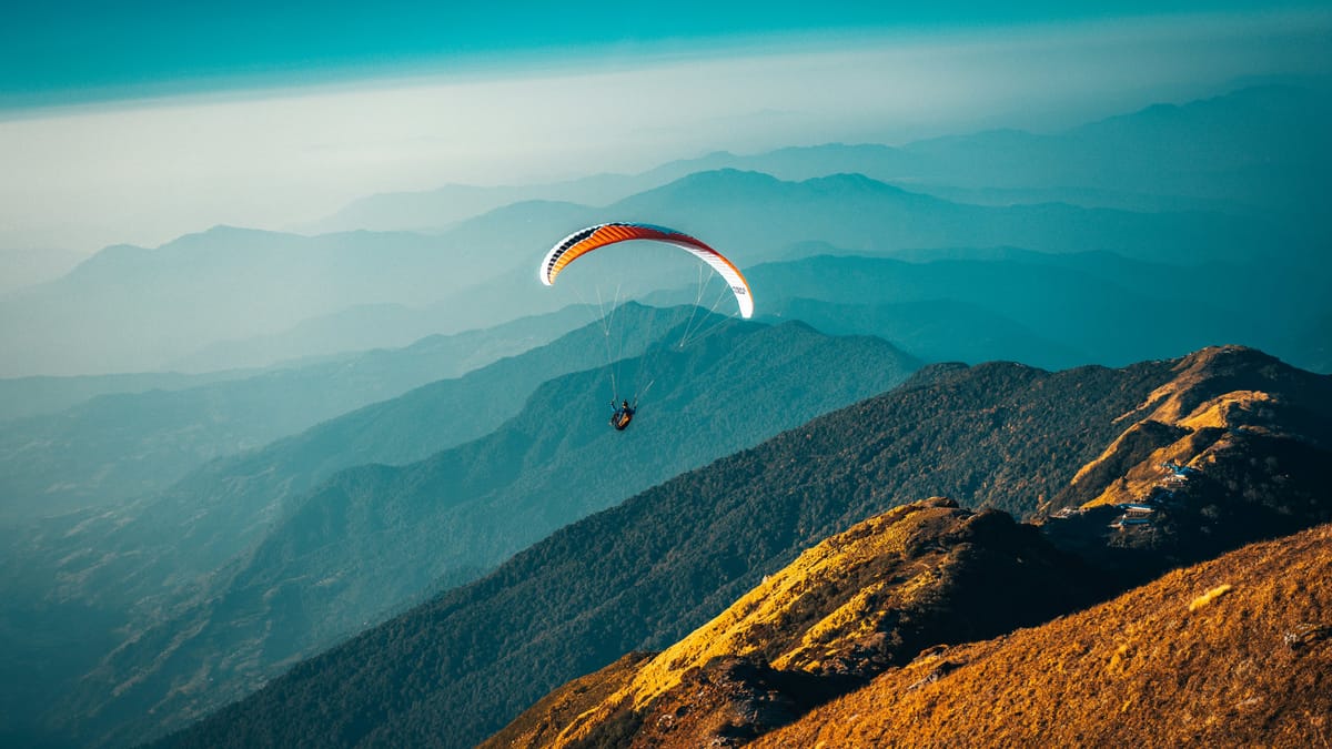 Ein Gleitschirmflieger schwebt hoch über einer weitläufigen Berglandschaft, deren Hügelketten in blaugrünen Schichten bis zum Horizont verlaufen.