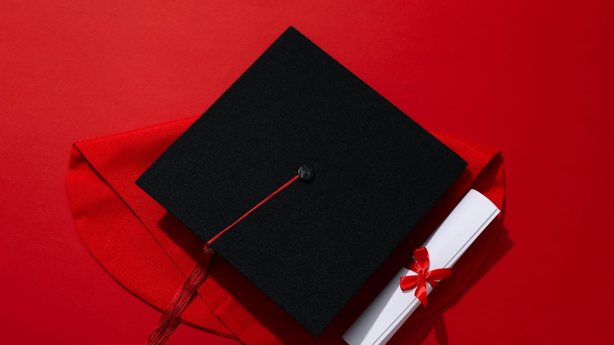 Ein schwarzer Absolventenhut mit roter Quaste liegt auf rotem Stoff, daneben eine zusammengerollte Urkunde mit roter Schleife.