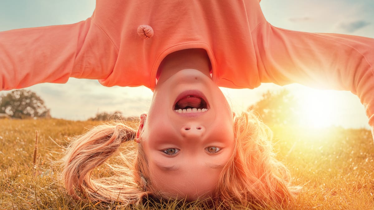 Ein lachendes Mädchen macht einen Kopfstand auf einer Wiese. Im Hintergrund scheint die Sonne und taucht die Szenerie in ein warmes, orangenes Licht.